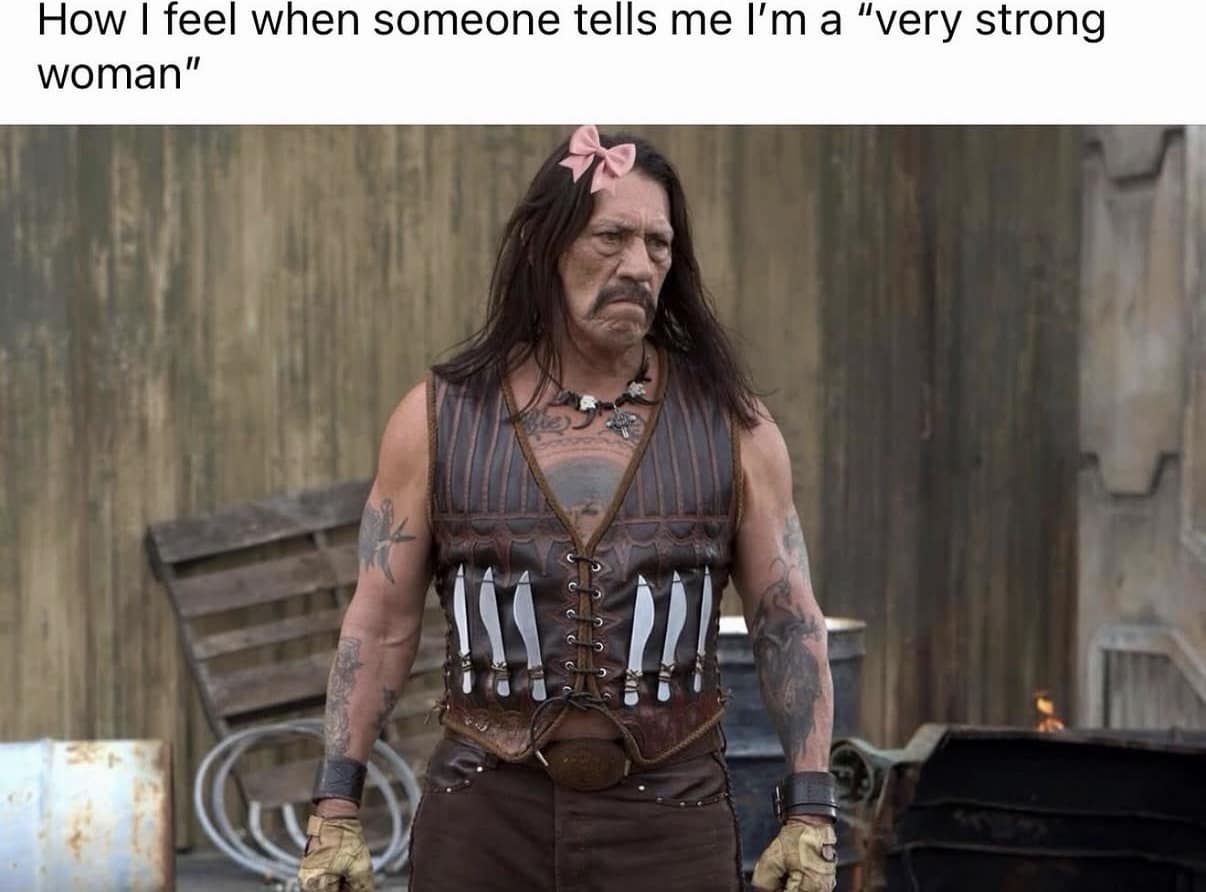 A funny meme featuring Danny Trejo in full "Machete" gear with a leather vest of knives, but sporting a tiny pink bow in his hair to represent the feeling of being called a "very strong woman."