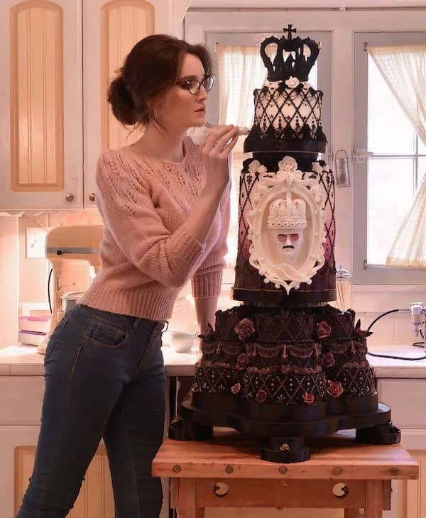 A woman in a kitchen finishing an elaborate, multi-tiered black and white gothic-style cake with crowns.