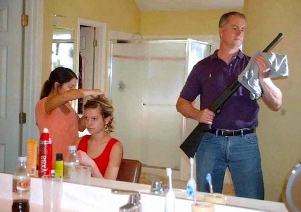 A safety third nightmare showing a man cleaning a shotgun in a cramped bathroom while two women stand inches away, mirror-adjacent, getting ready for an event.