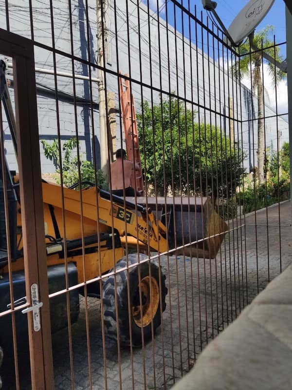 A multi-layered safety third failure showing a shirtless man on a ladder that is balanced inside the raised bucket of an orange excavator to reach overhead utility lines.