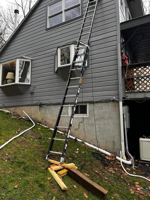 safety third masterpiece showing the base of a long extension ladder balanced on a wobbly, uneven pile of scrap wood on a steep grassy slope.