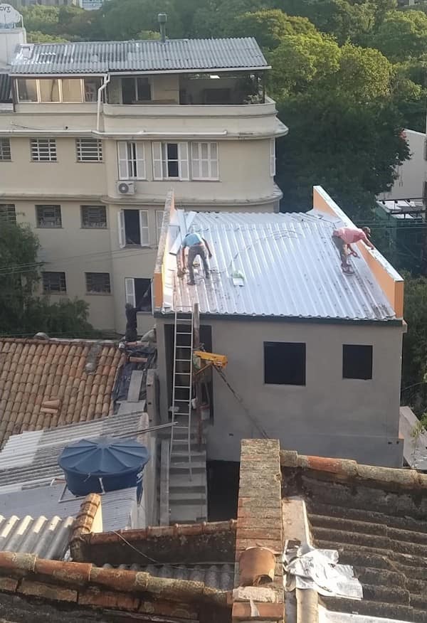 Two roofers working on a corrugated metal roof with a massive vertical drop and zero visible fall protection or harnesses.