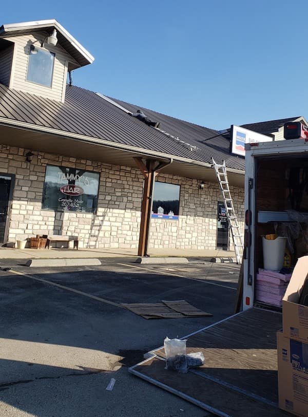 A DIY roofing setup where an extension ladder is propped against a storefront, held in place only by the open back doors of a white work van.
