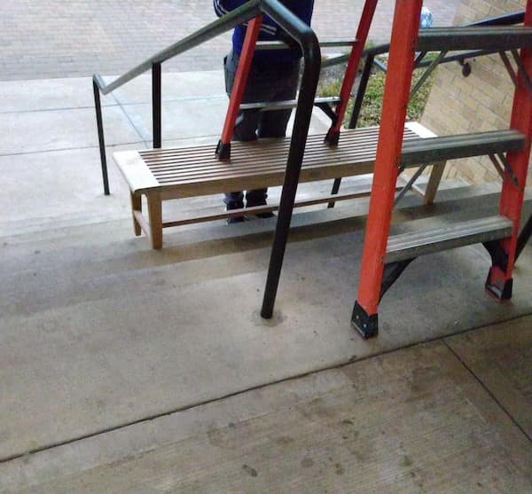 A safety third classic featuring a red step-ladder balanced on top of a wooden bench, which is itself balanced across three concrete stairs.