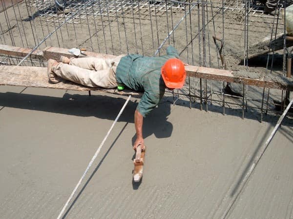 A construction worker lying face-down on a single wooden plank over a massive pit of wet concrete, using his hands to smooth the surface.