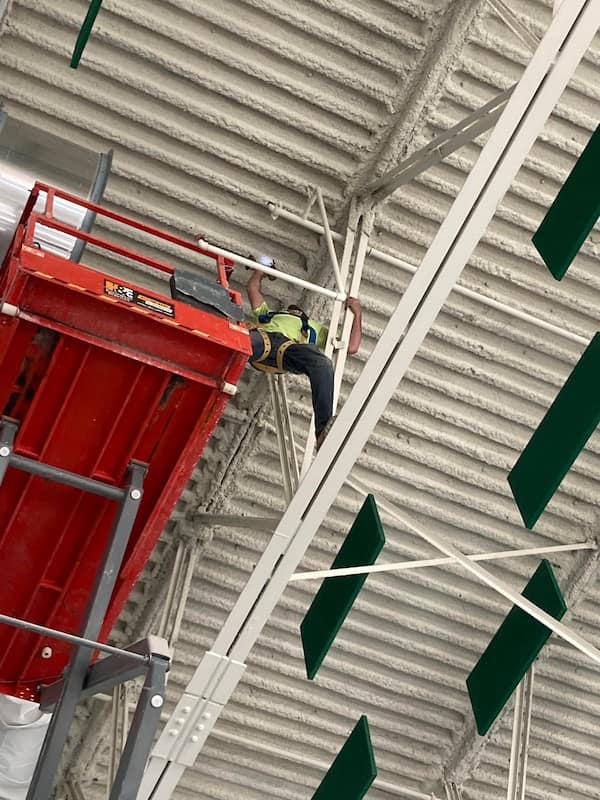 A worker wearing a harness but choosing to climb on top of the red scissor lift’s handrails to reach a high ceiling beam.