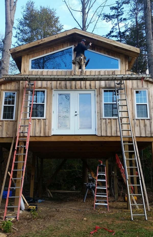 A precarious safety third construction setup where a man stands on a ladder laid flat across two vertical ladders to work on the upper windows of a house.