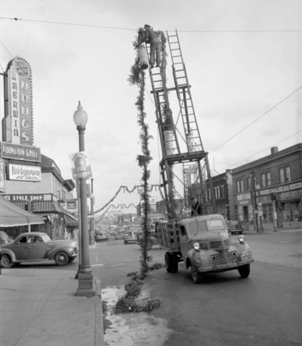 A vintage black and white safety third photo showing a utility worker on a tall, makeshift wooden scaffold built on the back of a moving truck to fix overhead wires.