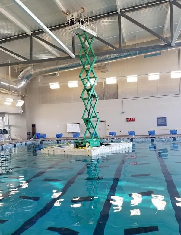 A breathtaking safety third moment featuring a fully extended green scissor lift balanced on a small floating platform in the center of an indoor swimming pool.