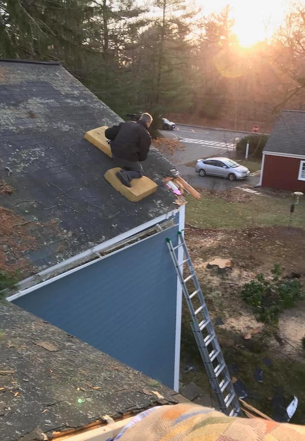A DIY safety third roofing project where a man kneels on loose foam blocks on a steep, mossy shingle roof while repairing a damaged corner.