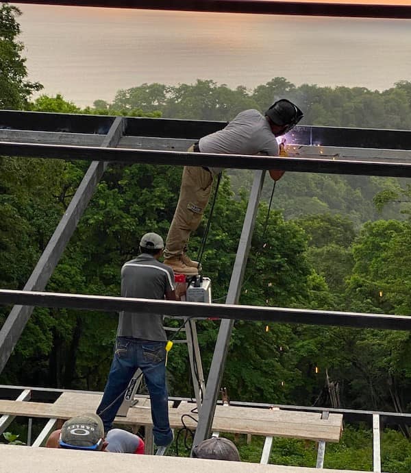 A high-stakes safety third welding operation showing one man standing on the very top step of a ladder positioned on a temporary wooden plank high above a forest.