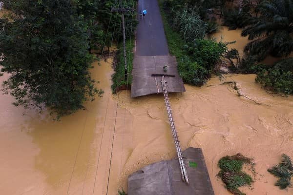 A terrifyingly thin safety third bridge made of lashed-together logs crossing a wide, churning brown floodwater current.