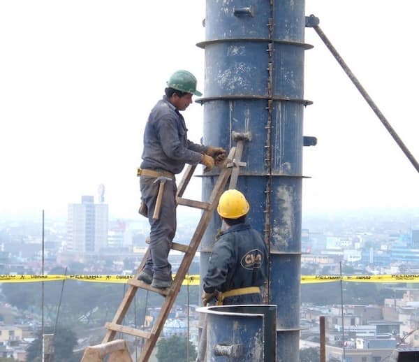 A vertical safety third challenge where a construction worker stands high on a wooden ladder propped against a large blue industrial pipe without visible harnesses.