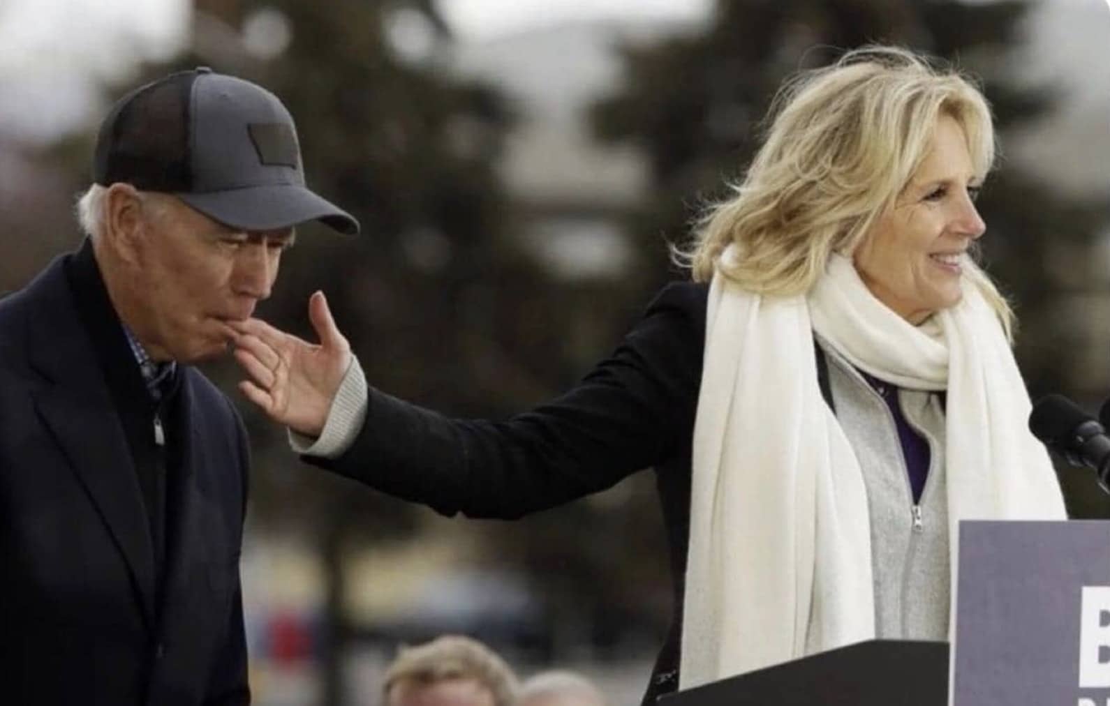 Jill Biden smiling at a podium while reaching back to touch Joe Biden's face.
