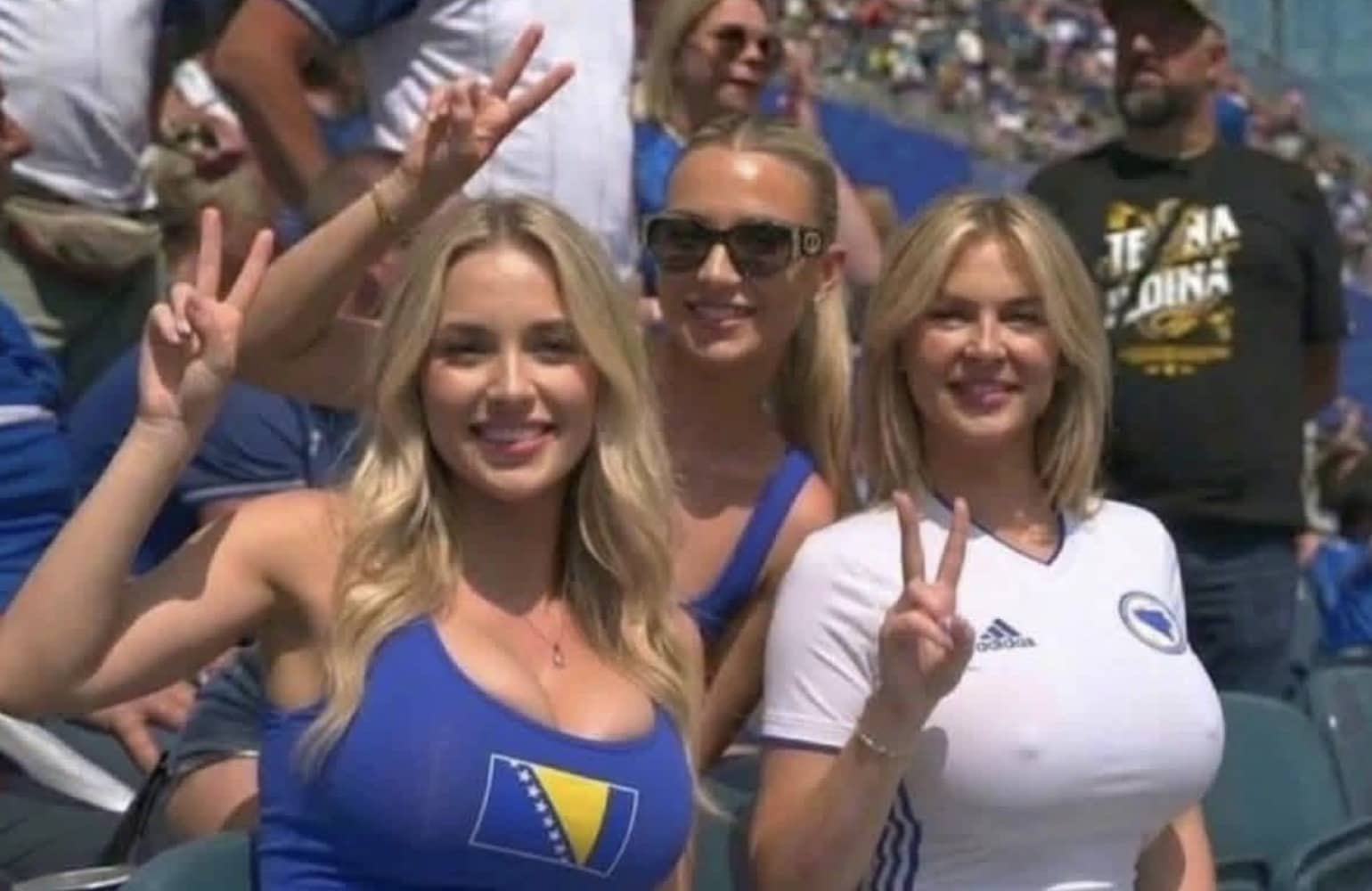 Memorable blonde female soccer fans smiling and posing with peace signs at a sports stadium.