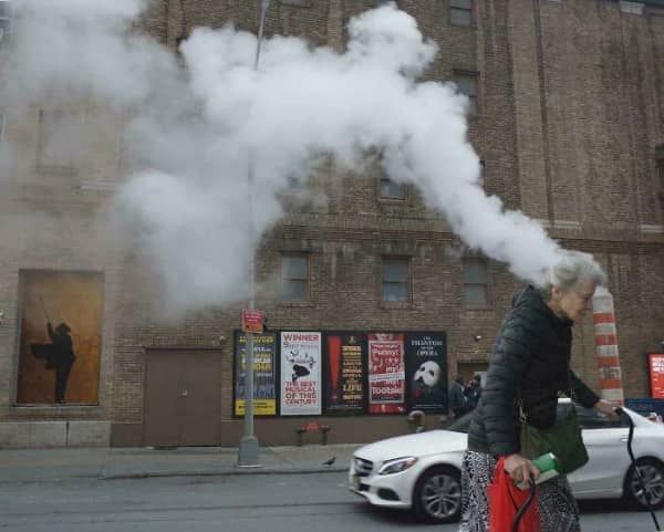 An elderly woman walking past a city building where background exhaust steam appears to exit her head.