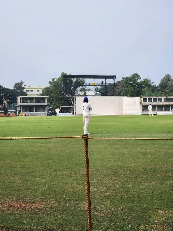 A cricket player standing far away on a pitch directly behind a fence post, looking tiny.