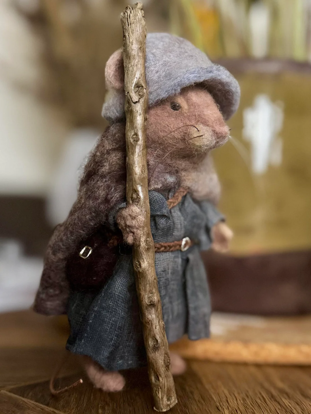 Small, adventurous felt mouse dressed in a traveler's cloak and pointed blue hat, gripping a tiny natural wooden staff in an example of narrative-driven funny felt art.