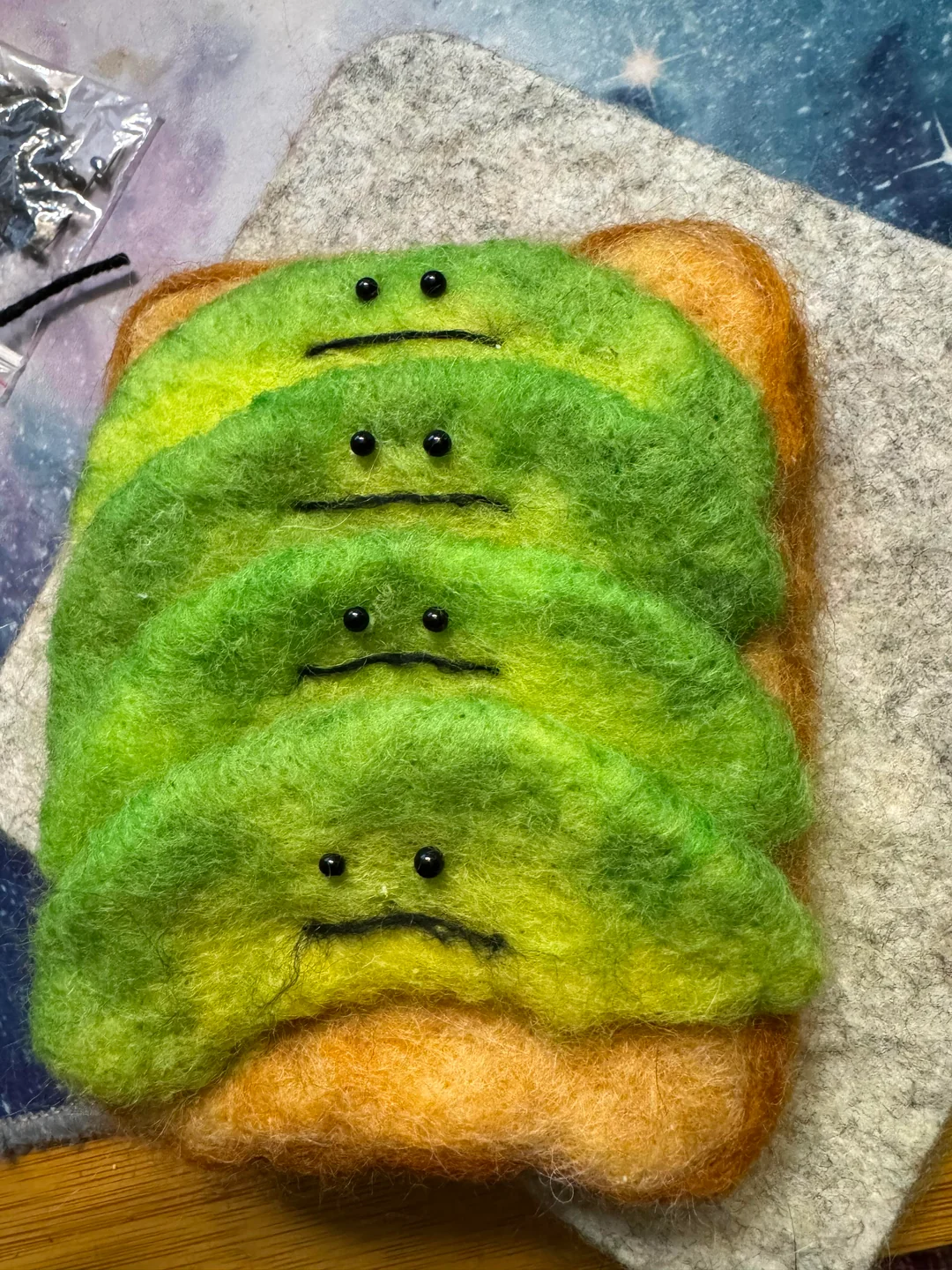 A thick slice of needle-felted wool toast topped with four stacked green avocado ridges, each given a pair of tiny black bead eyes and a hand-stitched sad, downturned mouth for an existential twist on funny felt art.