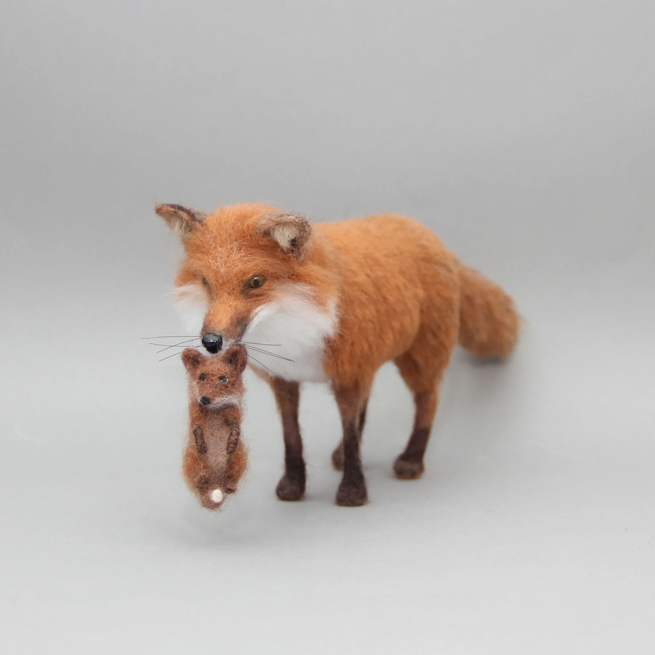 An astonishingly life-like, masterfully detailed needle-felted red fox figurine standing on a light grey background while gently carrying a tiny, matching realistic fox kit by its scruff, showcasing the absolute pinnacle of funny felt art.