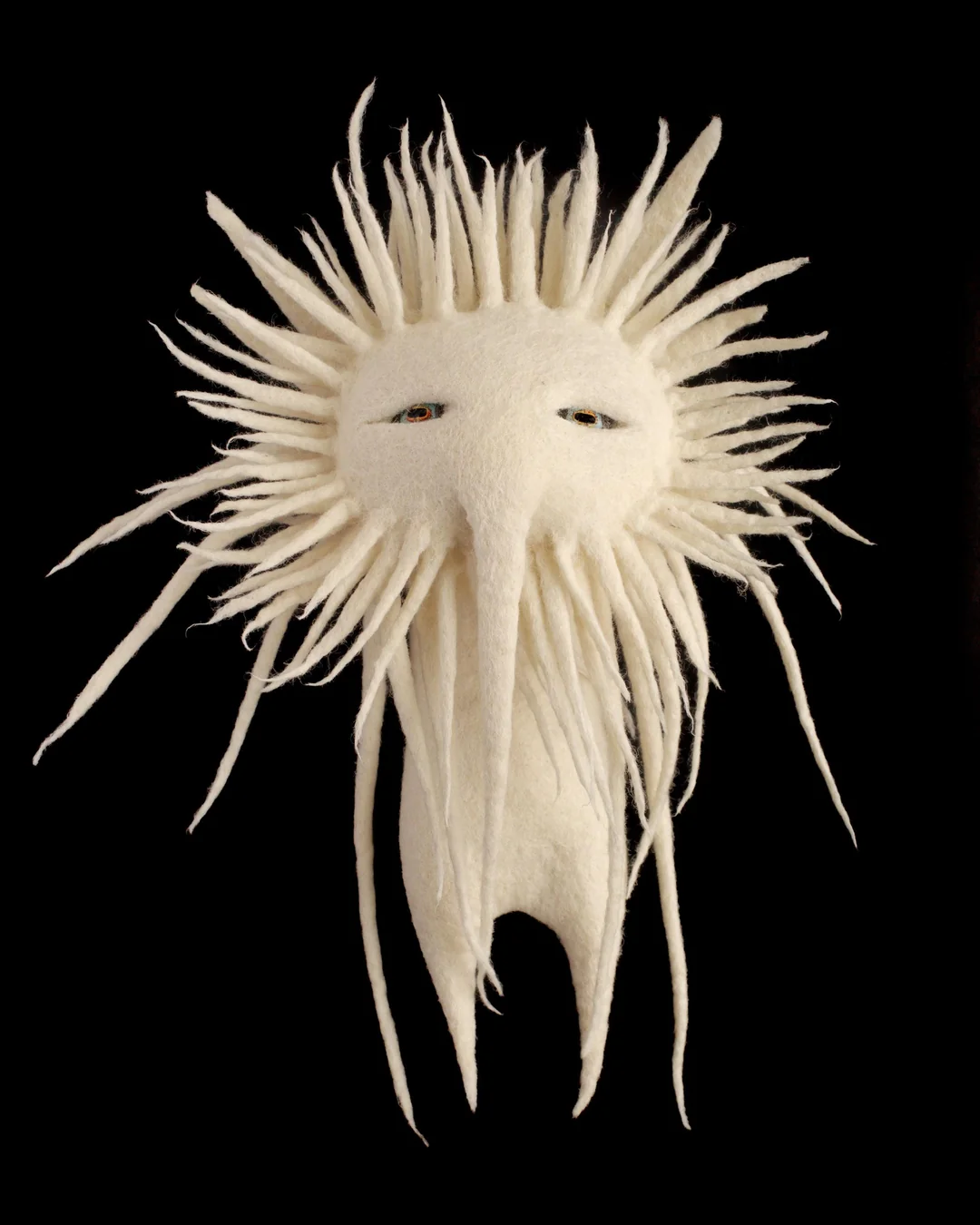A deeply surreal and eerie white wool cryptid doll featuring a giant circular face surrounded by sharp, sun-like spikes, narrow half-closed orange eyes, and a single elongated pointed nose beak extending down past its thin legs in an example of cursed crafts.
