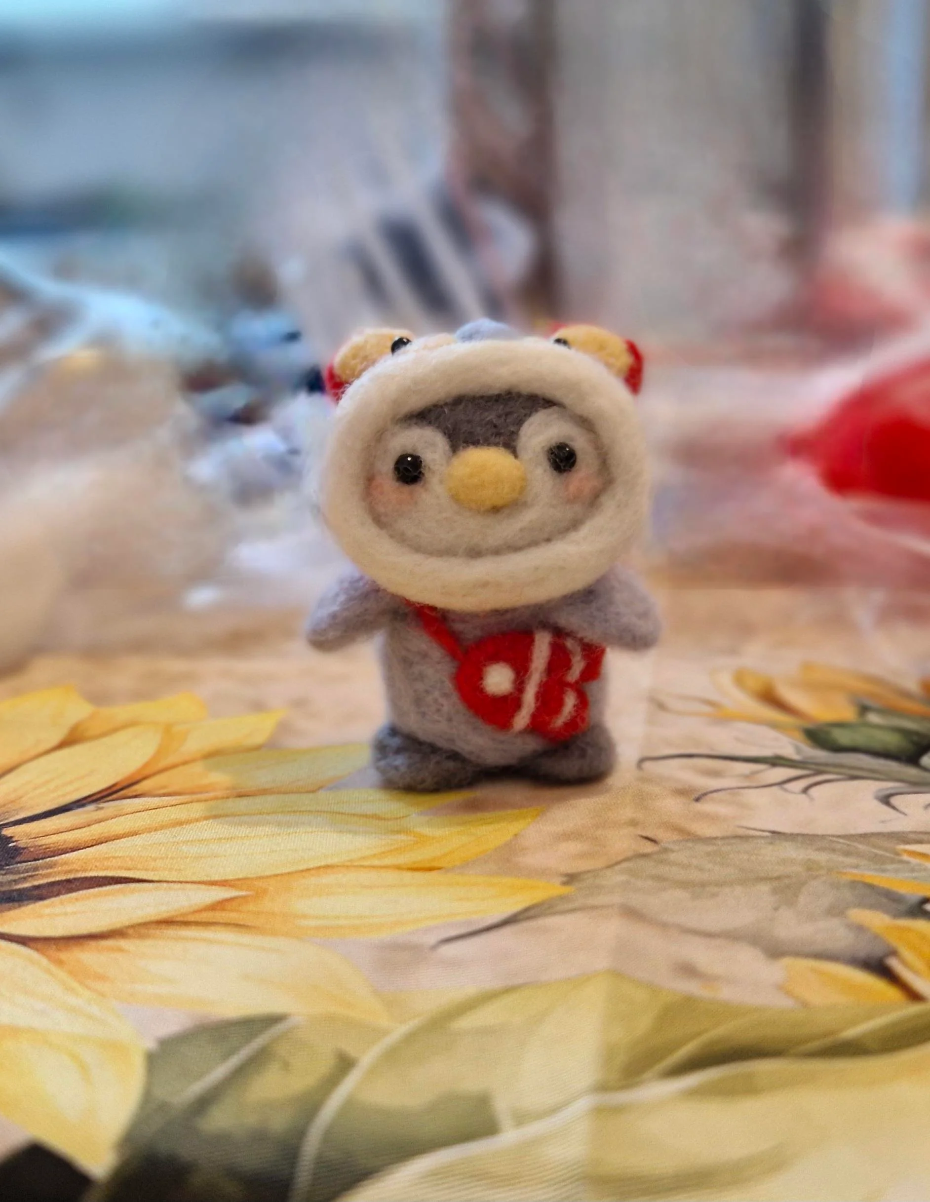 An ultra-cute, miniature grey felted penguin doll wearing a plush white winter hood with little teddy bear ears and sporting a tiny red satchel bag with a white heart accent, representing pristine funny felt art.