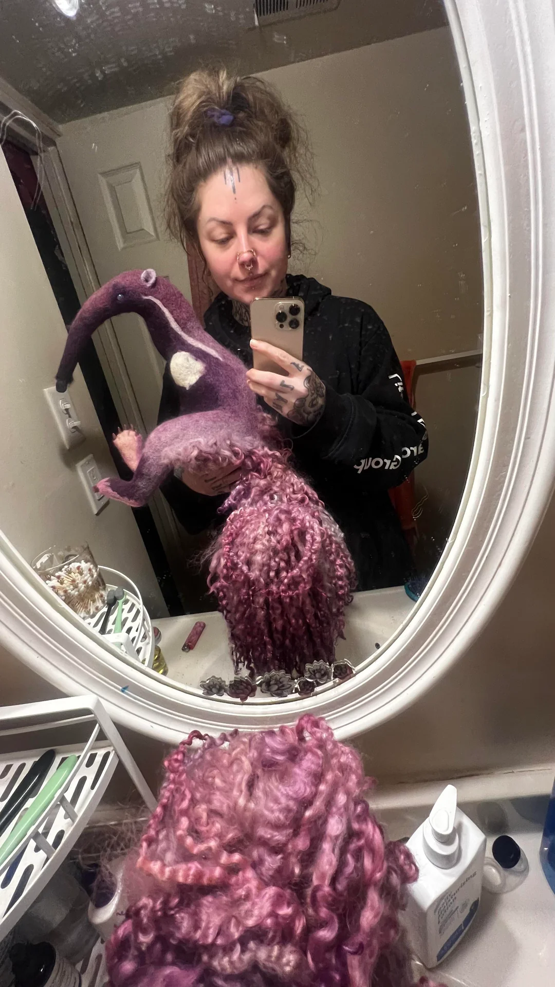 Bathroom mirror selfie of a woman holding a large, custom-sculpted purple felt anteater plush featuring an incredibly dense, flowing mane made from long, curly loops of pink and lavender dyed sheep wool, highlighting an artistic masterpiece of funny felt art.