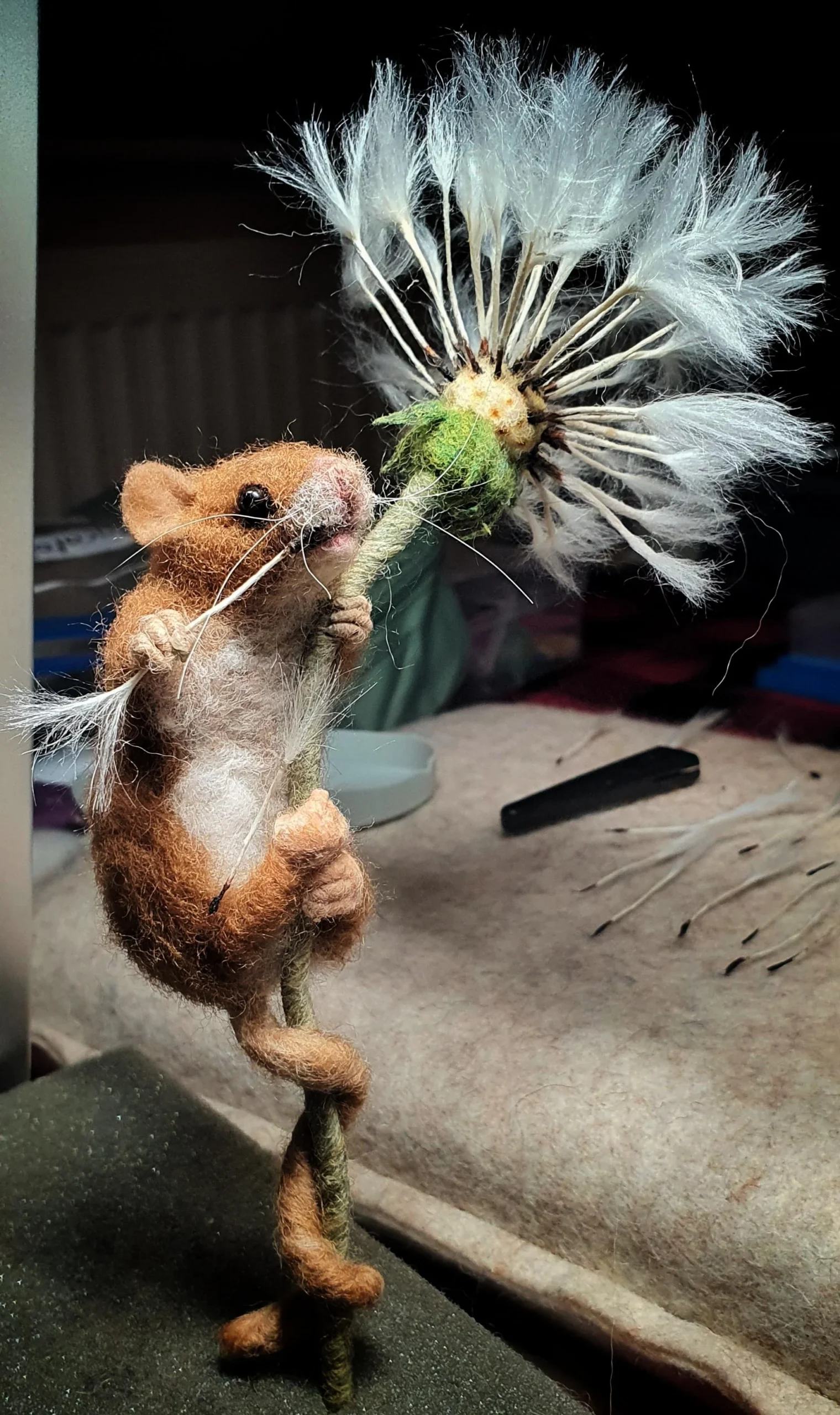 Exquisitely detailed needle-felted field mouse wrapping its tail and paws around a dandelion stem with realistic white seed puffs, displaying whimsical funny felt art.