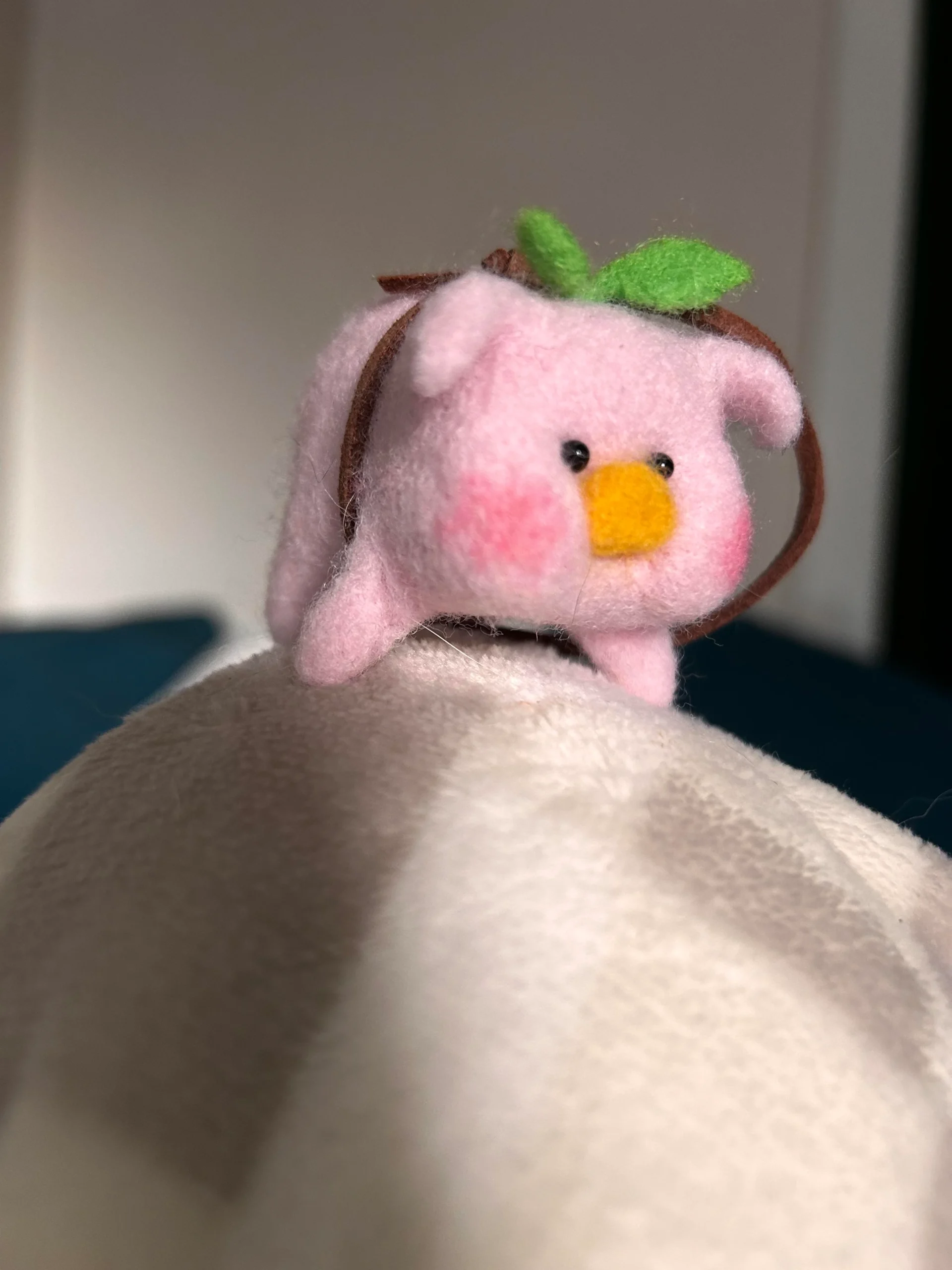 Chubby, miniature pink felt piglet creature with rosy cheeks and a green sprout on its head, resting on a plush toy as a piece of adorable funny felt art.
