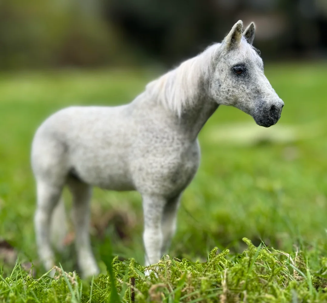 Strikingly realistic dapple-gray felted horse figurine standing upright in vibrant green mossy grass, showing a master-level transition away from cursed crafts.