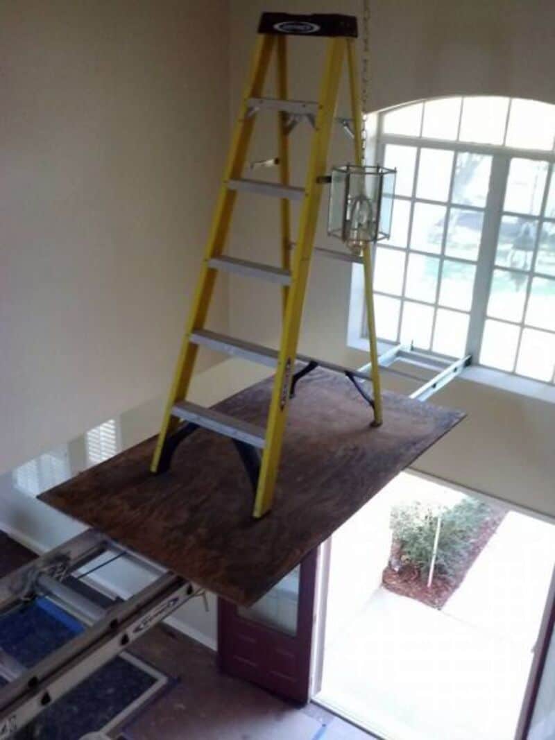 Unsafe construction photo showing a step ladder balanced precariously on plywood between two extension ladders.