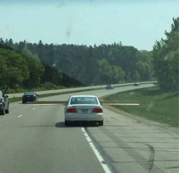 A white sedan driving down the highway carrying a long piece of lumber horizontally.