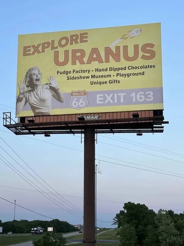 Massive yellow roadside billboard along Route 66 reading explore Uranus fudge factory funny meme.