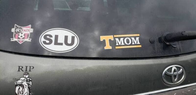 Automobile bumper with various stickers including university logos and a memorial motorcycle decal.