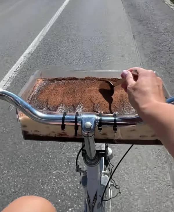 First person view of a bicycle handlebar holding a large glass tray filled with tiramisu dessert.