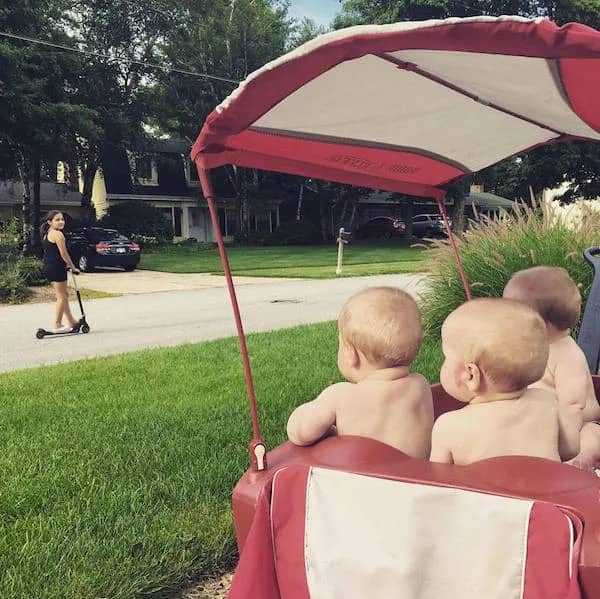 Three young babies in a red wagon watching a young girl ride a scooter.