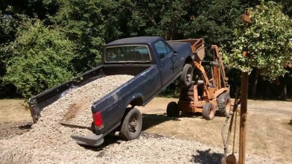 A blue pickup truck being tilted upwards by a yellow forklift to unload gravel.