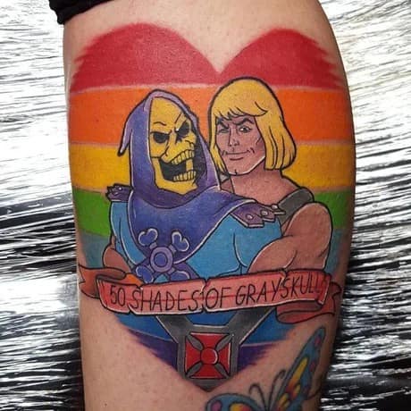 A vibrant skin tattoo showcased as a pop-culture pride meme, displaying a cartoon illustration of Skeletor and He-Man embracing inside a large rainbow-colored heart above a decorative banner reading "50 Shades of Grayskull."