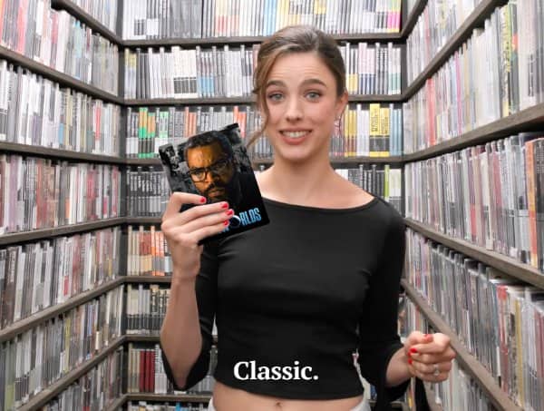 A young woman holding up a movie case in front of extensive shelves filled with media.