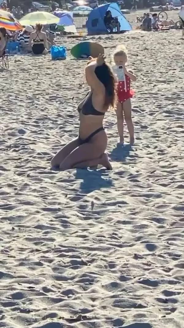 Young toddler holding a smartphone to photograph a woman posing on a sandy beach.