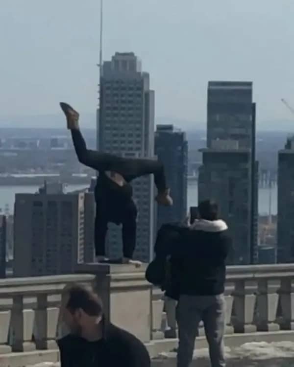 Person doing an extreme handstand on a high skyscraper stone ledge overlooking a city.