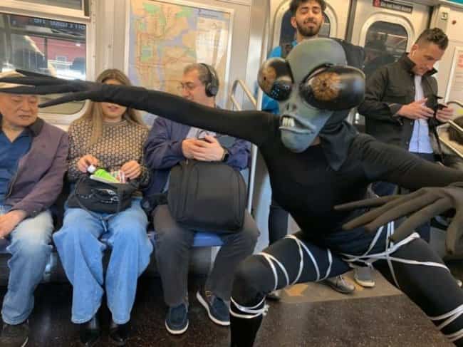 A person dressed as a bug alien posing dramatically inside a busy subway car for public transit memes.