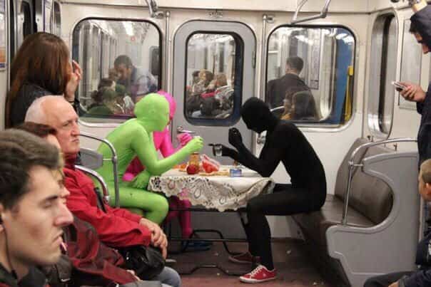 People in colorful morphsuits eating at a table inside a crowded subway car for public transit memes.