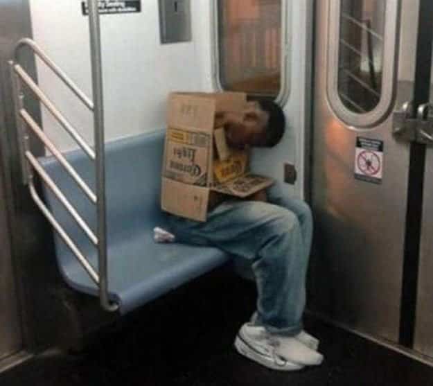 A sleeping train passenger hiding his head inside a cardboard Corona beer box for public transit memes.