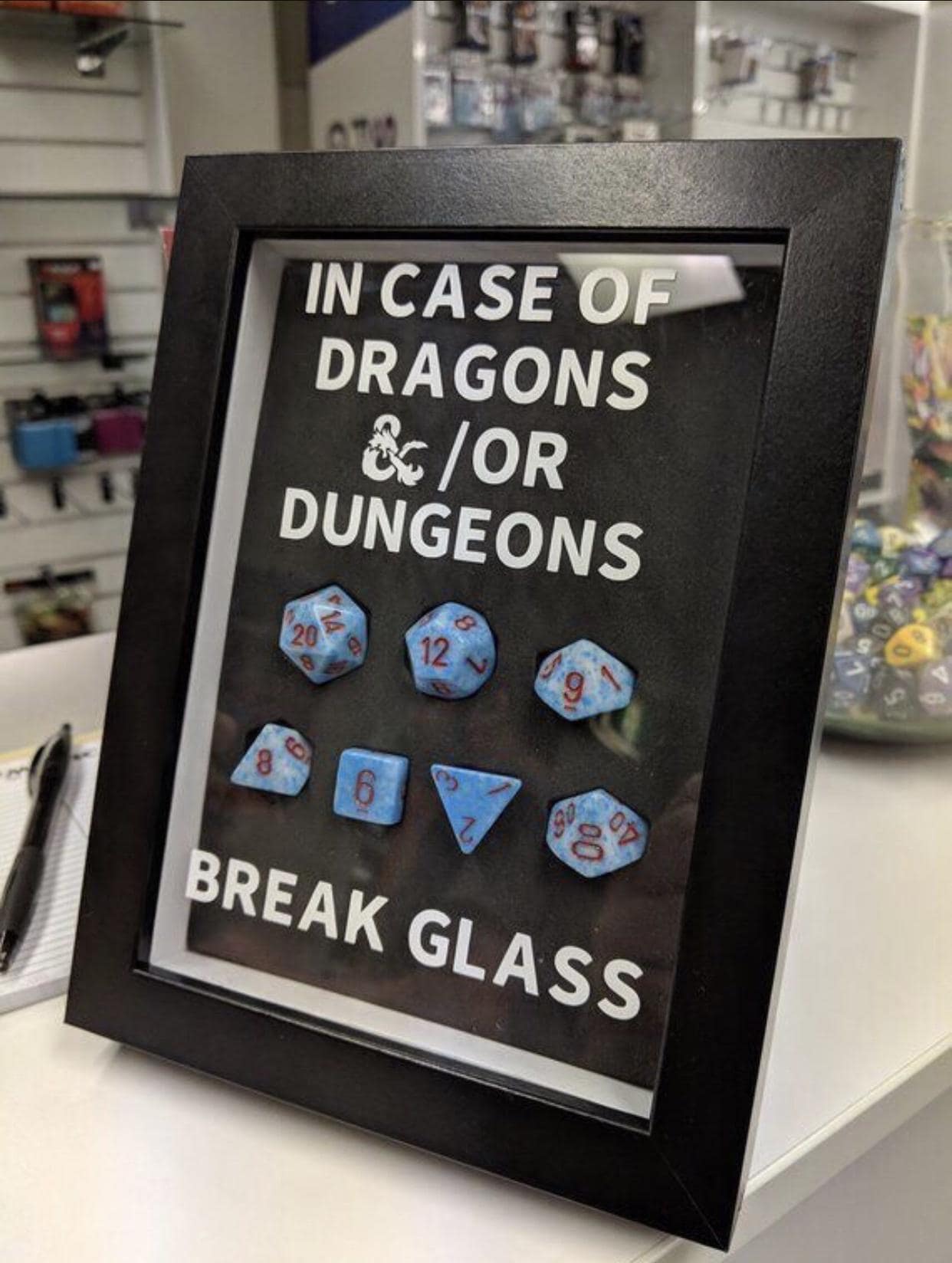 A brilliant d&d meme features a black emergency shadowbox frame displayed on a retail store counter. Bold white lettering on the glass reads, "IN CASE OF DRAGONS &/OR DUNGEONS BREAK GLASS," physically protecting a full set of seven light blue marble-textured polyhedral dice with sharp red numbers, including a prominent d20, d12, and d10.