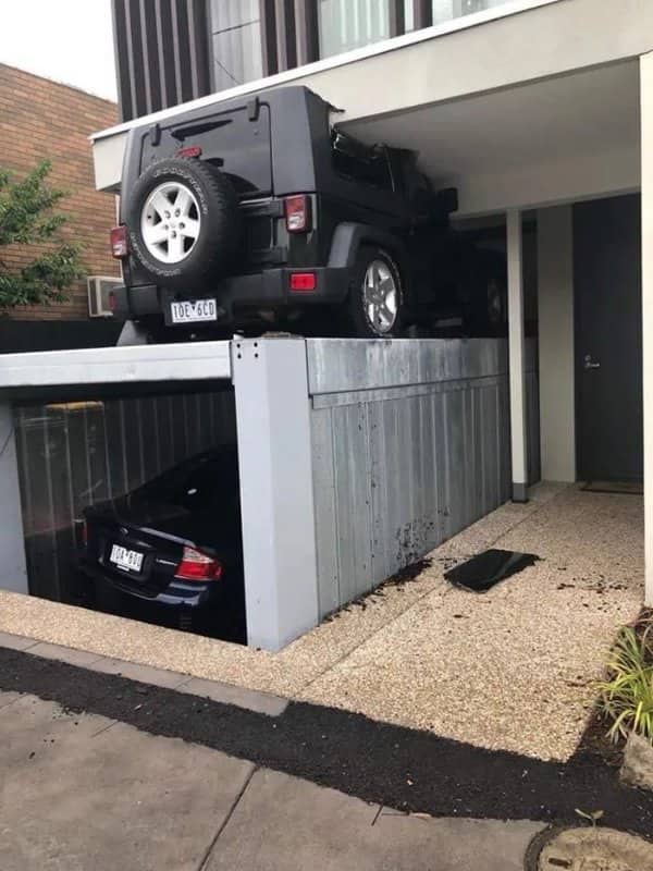 A black Jeep crushed under a concrete ceiling on a multi-level stacking garage lift.