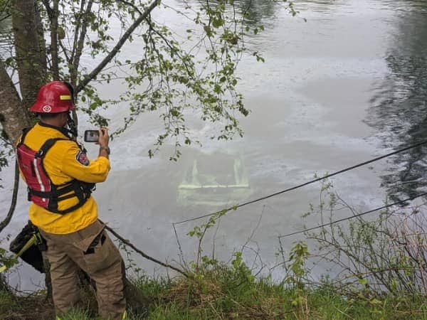 A firefighter taking a photo of a fully submerged car hidden just beneath river water.