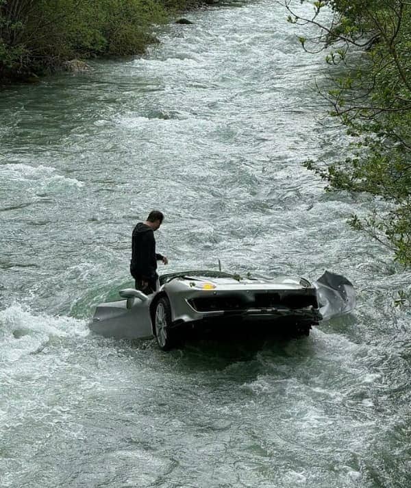 A luxury silver sports car floating down a rapidly flowing rocky river stream with a bystander.