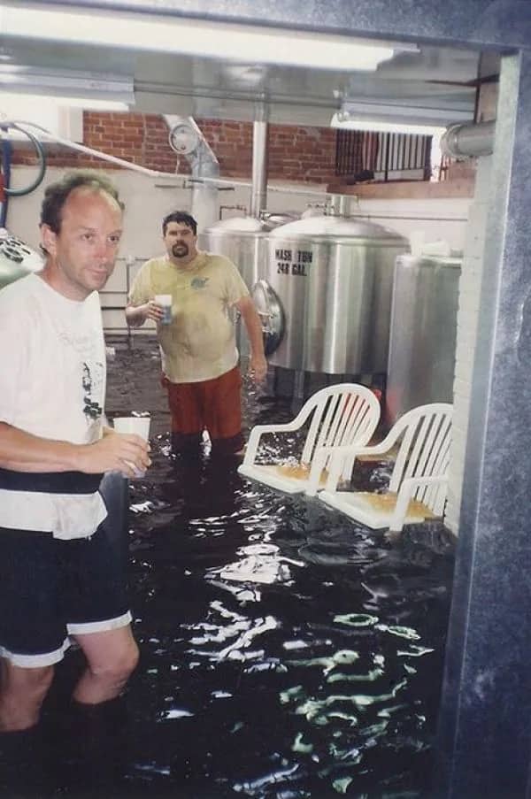 Two men standing knee-deep in dark flooded water inside a brewery next to empty plastic chairs.