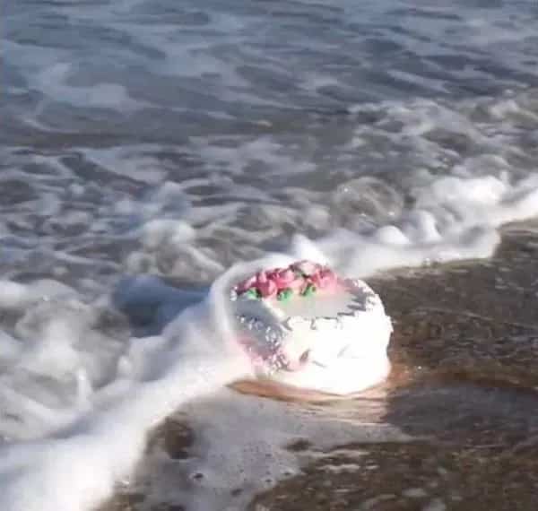 Poetic or symbolic funny meme showing a beautiful pink frosted birthday cake being washed away by ocean waves.